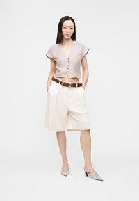 Μια γυναίκα στέκεται με τα χέρια στις τσέπες, φορώντας ένα λιλά κοντό πουκάμισο με κουμπιά, κρεμ σορτς μήκους έως το γόνατο με ψηλή μέση, καφέ ζώνη και μπεζ ψηλοτάκουνα.