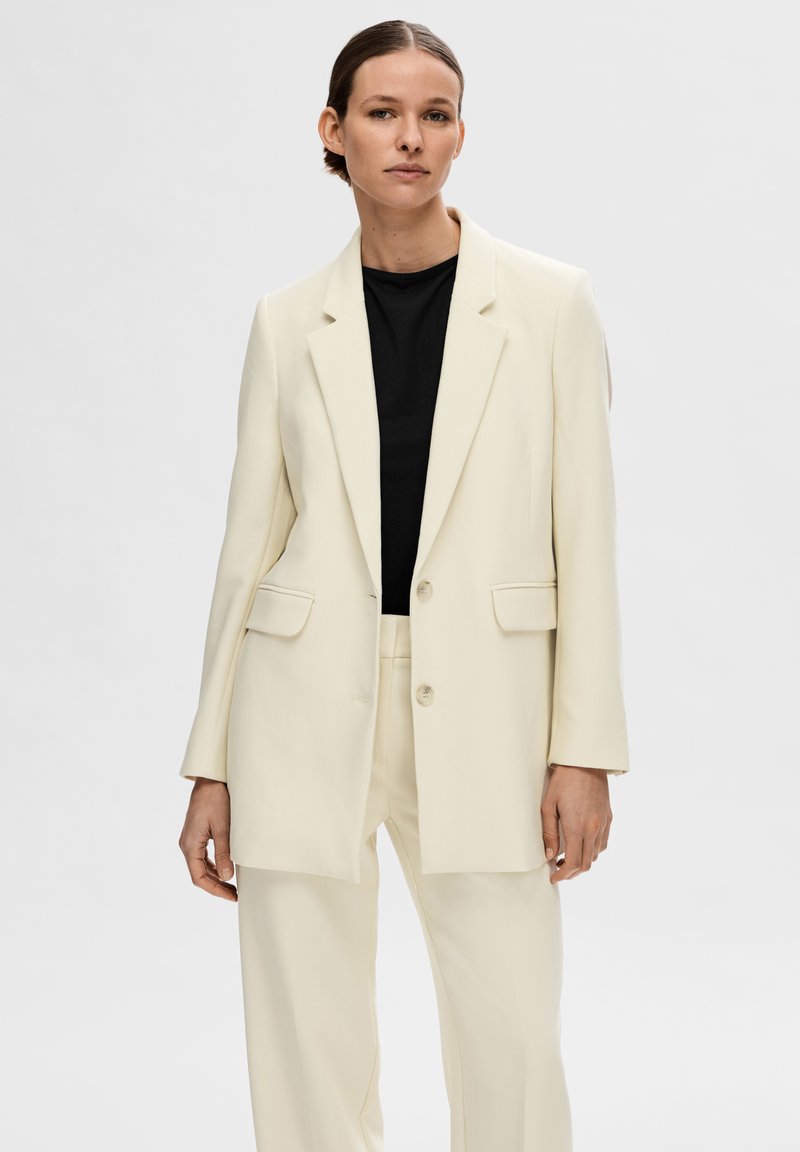 Cremefarbener oversized Blazer mit zwei Knöpfen, Reverskragen und Seitentaschen; kombiniert mit einem schwarzen Top in minimalistischer, glatter Ausführung.