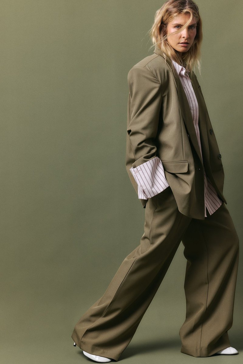 Olīvu zaļš pārsteidzīgs žakets un saskaņotas plaša griezuma bikses, stilizētas ar gaisīgu svītrains kreklu, kas piedāvā lielus manšetes un gludu auduma tekstūru.