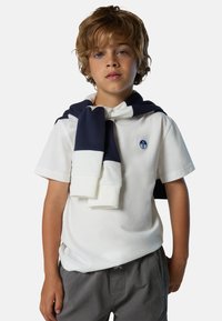 Weißes Poloshirt mit kurzen Ärmeln und Logo, kombiniert mit einem navyblauen und weißen Sweater, der um die Schultern gebunden ist, und grauen Shorts. Sichtbare Texturdetails des Stoffs.