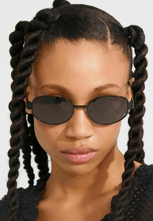 Crne ovalne sunčane naočale s toniranim staklima i tankim crnim okvirima. Subjekt ima teksturiranu, uvijenu kosu stiliziranu u više segmenata.