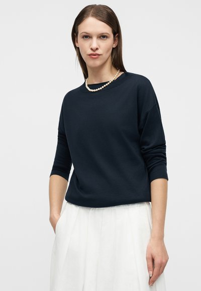 Γυναίκα με ίσια καστανά μαλλιά που φοράει μπλε μπλούζα μακρυμάνικη, λευκή φούστα και μαργαριταρένιο κολιέ, όρθια με το ένα χέρι στην τσέπη.