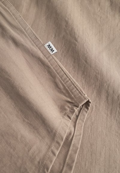 Tela beige con un sutil patrón texturizado, que presenta costuras cuidadosas y una pequeña etiqueta de marca blanca. Material suave y ligero.
