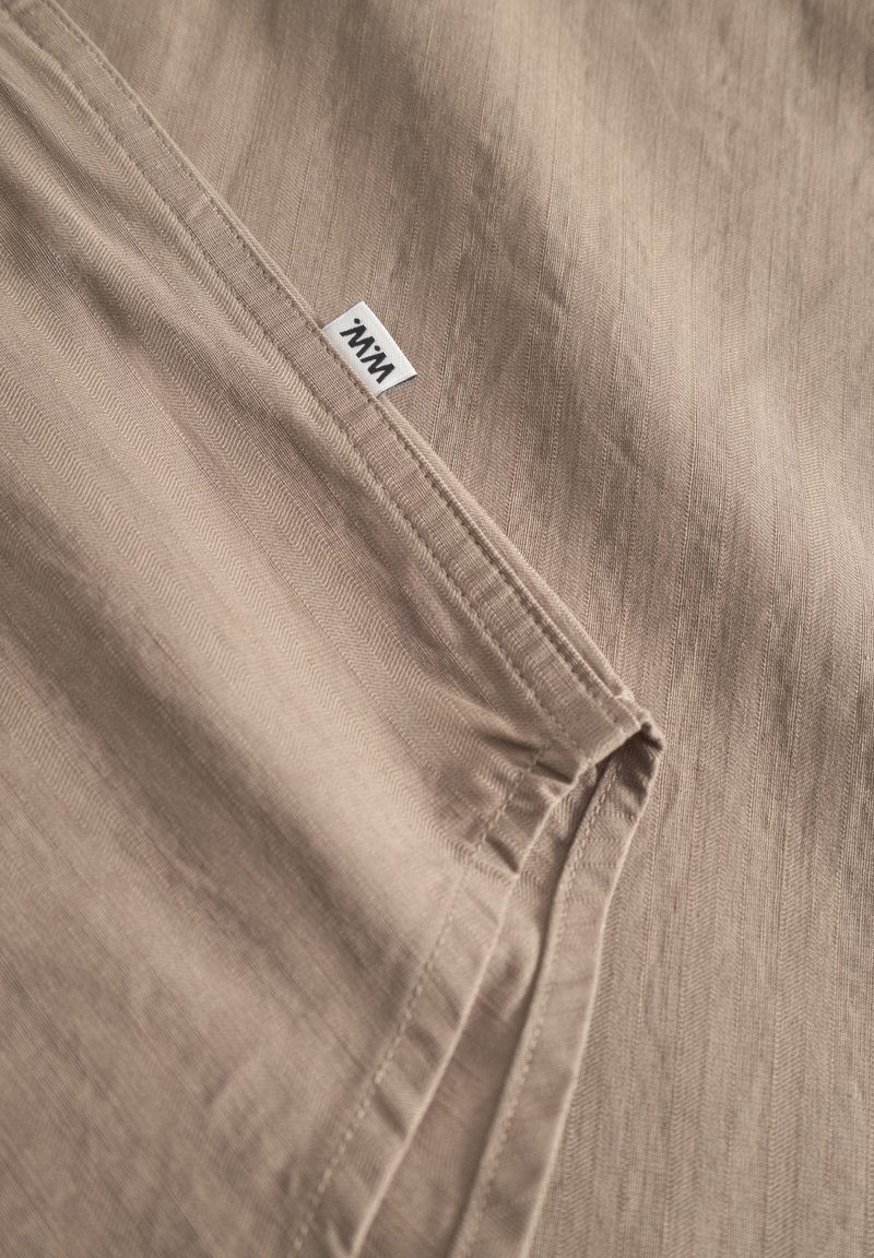 Tissu beige avec un motif texturé subtil, présentant des coutures soignées et une petite étiquette de marque blanche. Matériau lisse et léger.