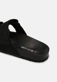 Sandales noires avec un lit de pied texturé, un design à lanières ouvertes et une semelle plate. Présente un logo visible sur le lit de pied et un matériau en daim doux.