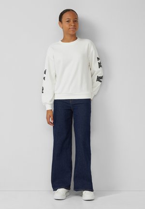 Sudadera blanca con acentos gráficos negros en las mangas, combinada con jeans de pierna ancha azul oscuro y zapatillas blancas. Textura suave.