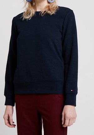 Osoba ubrana w granatową, długorękawową bluzę z małym logo na mankiecie rękawa oraz ciemnoczerwone spodnie, stojąca na jednolitym tle.