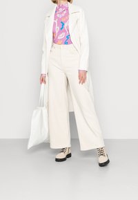 Beige weite Hose, pink gemusterte Bluse, weiße Jacke und beige Stiefel mit schwarzen Sohlen, hält eine weiße Tragetasche, vor einem grauen Hintergrund.