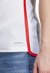 Valkoinen urheilupaita punaisilla sivuvarusteilla, teksturoitu kangas, jossa on "AEROREADY" -merkki helman kohdalla. Slim fit -muotoilu, esitetty yhdessä farkkujen kanssa.