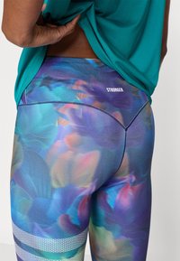 Κολάν με ζωηρό αφηρημένο σχέδιο σε αποχρώσεις του μπλε, του μοβ και του ροζ.  Περιλαμβάνει λευκό λογότυπο "STRONGER" και υφασμένους τόνους.