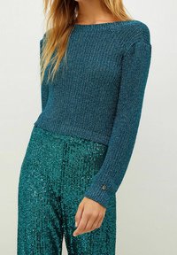 Woman wearing a blue knit long-sleeve sweater and teal sequined pants, with relaxed hand by her side.