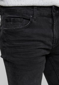 Černé džíny z denimu s úzkým střihem, které mají přední kapsu, zapínání na knoflík a jemné známky opotřebení na látce.