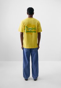 Camiseta de algodón amarilla con texto verde que dice "Hamelodi Sundowns" en la parte posterior. Combinada con unos vaqueros de denim azul holgados.