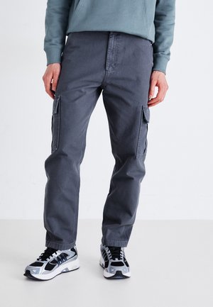 Pantaloni cargo grigi con due tasche laterali, chiusura con cerniera, tessuto resistente, gamba attillata, abbinati a scarpe da ginnastica nere e argento.
