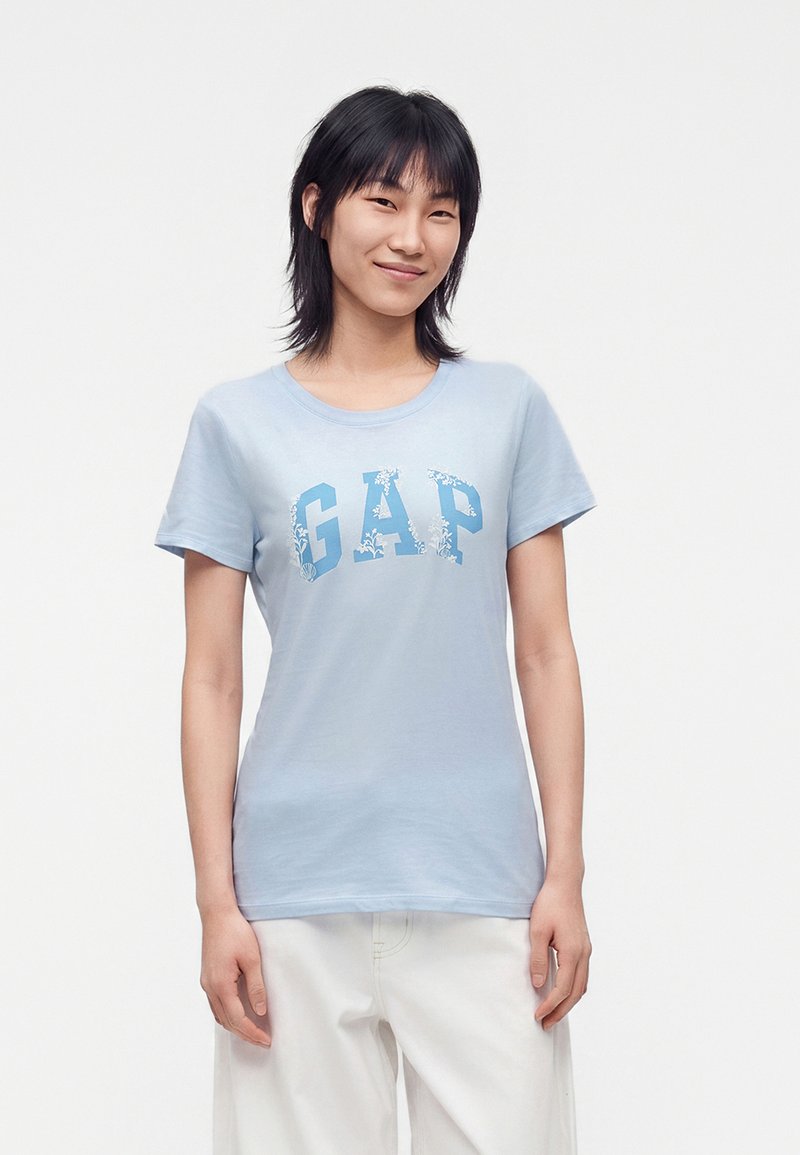 Personne aux cheveux noirs raides, portant un t-shirt GAP bleu clair avec lettrage floral et un pantalon blanc, debout devant un fond clair uni.