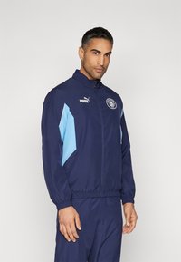 Mornarska jakna s kapuco z lahkimi modrimi stranskimi paneli, zadrgo spredaj, visoko ovratnico in elastičnimi manšetami, ki vsebuje logo Puma in emblem ekipe.
