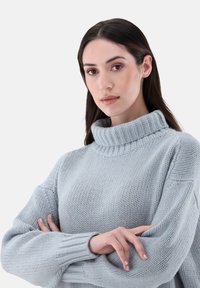 Hellblauer gestrickter Pullover mit hohen Rippkragen und locker sitzenden langen Ärmeln. Die Struktur ist dick und weich und zeigt subtile Stichmuster.