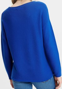 Femme portant un pull texturé bleu vif à manches longues et un jean bleu clair, vue de dos sur un fond uni.