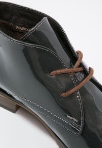 Chaussure en cuir verni gris foncé avec une finition lisse, lacets marron et surpiqûres blanches contrastantes. L'accent en bois sur le talon ajoute du détail.