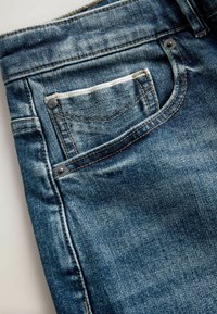 Denimjeans i blekt blått med en strukturerad yta; har en framficka med vit sömnad och silverfärgade detaljer.