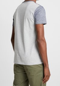 Hombre con una camiseta de dos tonos en gris de manga corta y pantalones verdes oliva, de espaldas a la cámara contra un fondo liso.