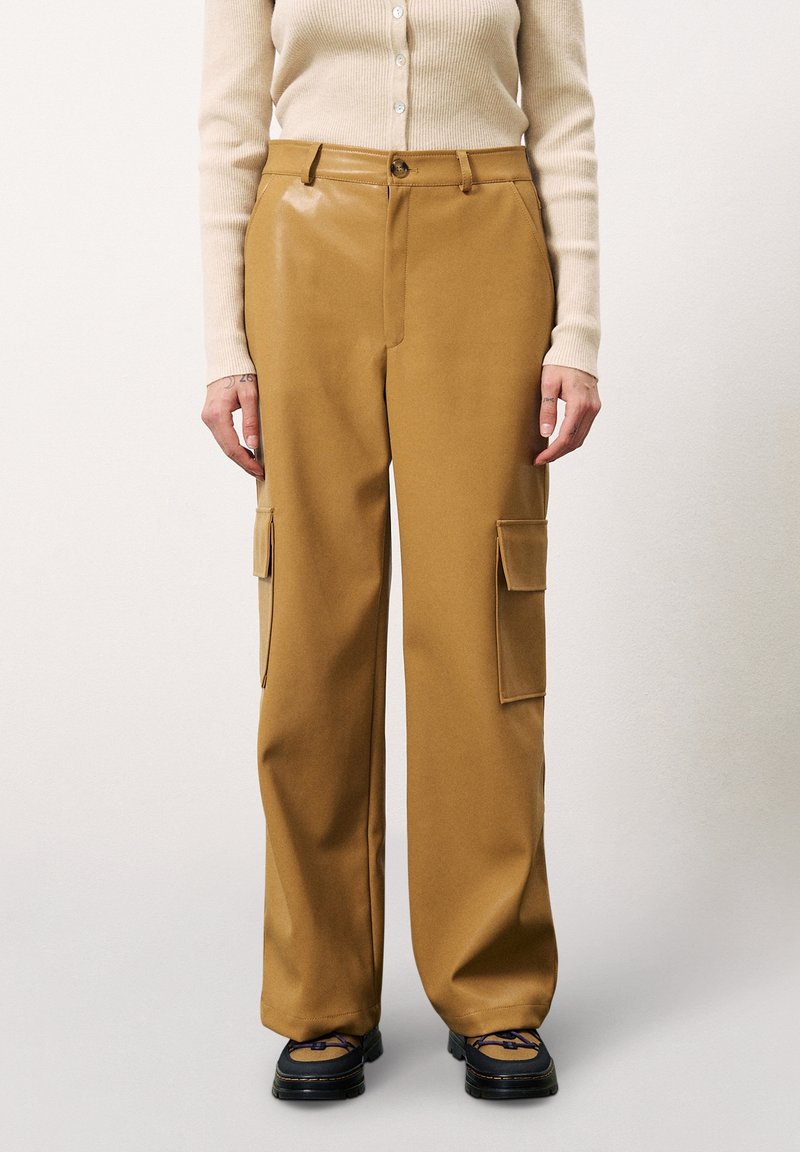 Personne portant un pantalon cargo large beige avec poches à rabats latérales, un haut côtelé crème à manches longues et des chaussures noires avec des lacets bleus.