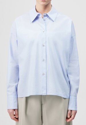 Vaaleansininen pitkähihaiset napitettava paita leveällä kauluksella, pidetty liehuvana beigejen housujen päällä, kuvassa henkilö niskasta lantioon asti.