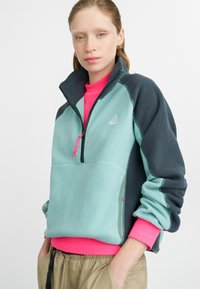 Fleece pulover z teal sprednjem delu, temno sivimi rokavi in rožnatim ovratnikom. Zadrga z rožnatimi vrvicami in detajl z logotipom.