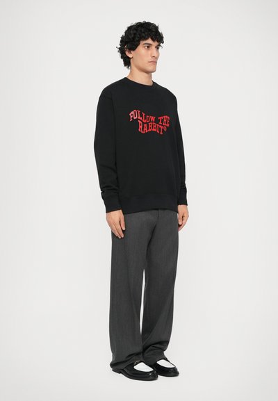Sort sweatshirt med rød tekst "FOLLOW THE RABBIT", parret med grå plisserede bukser og sorte sko med hvide detaljer. Model står sideværts.