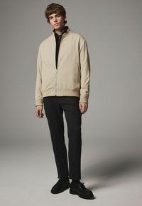Lys beige zip-jakke med ståkrave, parret med sorte bukser og sorte snørede sko. Glat stof og minimalistisk design.