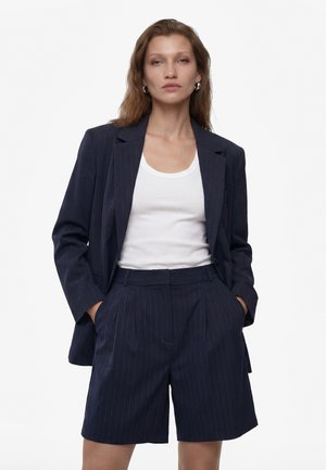 Kvinde iført navyblå skjortejakke med striber og matchende højtaljede shorts med hvid tanktop, hænderne i lommerne, mod hvid baggrund.