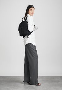 Μαύρος καμβάς σακίδιο με μπροστινή τσέπη και λογότυπο, κρατιέται από ένα άτομο που φορά λευκό πουκάμισο και γκρι παντελόνι, μπροστά από ουδέτερο φόντο.