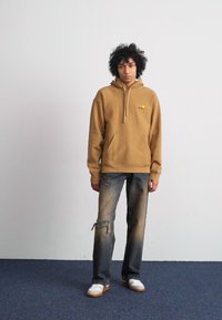 Brown hooded sweatshirt with kangaroo pocket, paired with distressed light blue jeans. White sneakers with light brown soles.