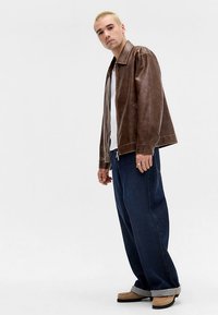 Brązowa skórzana kurtka o luźnym kroju, zestawiona z luźnymi ciemnymi dżinsami i beżowymi butami, na jednolitym białym tle.