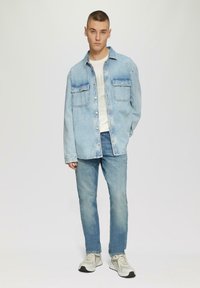 Jasnoniebieska koszula dżinsowa z długimi rękawami i kieszeniami na piersi, w połączeniu z jasnoniebieskimi dżinsami i szarymi trampkami. Pod spodem gładka biała koszulka.