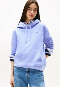 Fioletowa bluza z kapturem, wykonana z miękkiego materiału, z kieszenią kangurką, regulowanym kapturem i paskami na mankietach. Małe logo nad kieszenią.