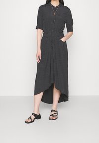 Kobieta w czarnej sukience midi w białe kropki, z rękawami do łokcia, guziki z przodu i czarne sandały na niskim obcasie z paskami.