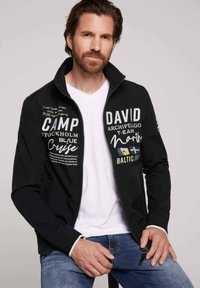 Sort zip-jakke med hvid tekst og logoer på forsiden. Har en ståkrave og ribbede ærmer, parret med en hvid V-hals t-shirt under.