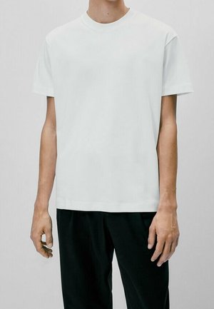 Personne portant un t-shirt blanc uni à manches courtes et un pantalon noir, debout devant un fond gris clair uni.