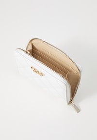 Portefeuille matelassé blanc avec quincaillerie dorée, fermeture éclair, et un intérieur beige comprenant des emplacements pour cartes et une petite poche.