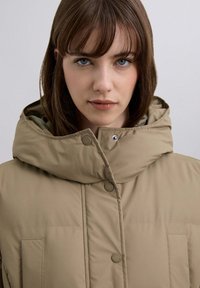 Chaqueta acolchada beige con cuello alto, grandes botones y bolsillos delanteros, que muestra una textura lisa y acolchada y un diseño minimalista.