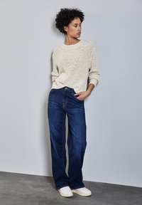 Maglione beige lavorato a maglia con maniche lunghe, abbinato a jeans in denim blu a gamba larga e sneakers bianche su uno sfondo neutro.
