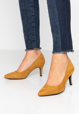 Brune ruskind høje hæle med spids tå og 7 cm stilethæl, kombineret med mørke denimjeans med frynset kant.