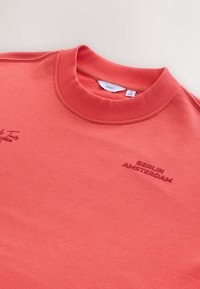 Felpa color corallo con scollo a coste. Presenta testo ricamato "BERLINO" e "AMSTERDAM" in corallo scuro, tessuto morbido e testurizzato.