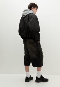 Čierna bomber bunda cez sivú mikinu, doplnená tmavými denimovými šortkami. Čierne tenisky dotvárajú outfit. Pozadie je jednoduché biele.