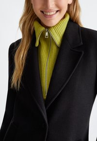 Cappotto di lana nera su un dolcevita a coste con zip di colore verde brillante. Dettagli in argento sulla zip molto evidenti e design della collo strutturato.