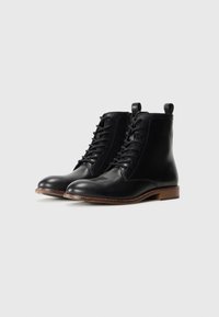 Botas de cuero negro hasta el tobillo con cordones, cremallera lateral, tiradores y tacones bajos marrones sobre fondo blanco liso.