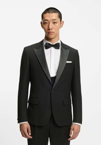 Giovane uomo con smoking nero con rever in raso, camicia bianca, papillon nero e fazzoletto bianco in tasca, su uno sfondo bianco semplice.