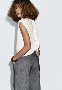 White sleeveless top with a back slit, paired with loose gray trousers featuring a textured finish and rear pockets.