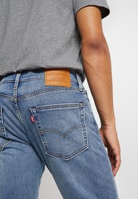 Jeans de mezclilla azul con una etiqueta de cuero en la cintura. Entre sus características se incluye un bolsillo trasero con costura curva y un diseño de corte ajustado.